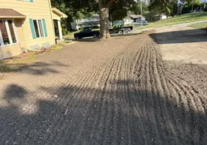 Freshly graded gravel driveway pad in a residential front yard with tine marks from grading, a house and a parked truck with utility trailer in the background