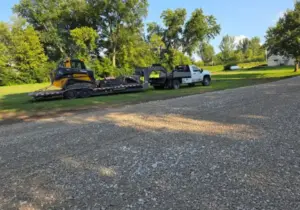 John Deere compact track loader loaded on a trailer being towed by a flatbed truck, parked on a gravel driveway in a grassy yard