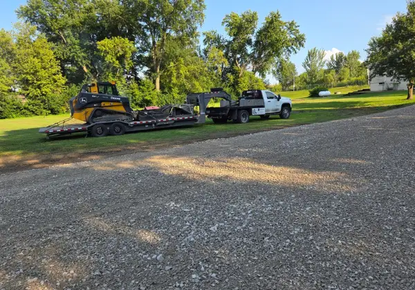 John Deere compact track loader loaded on a trailer being towed by a flatbed truck, parked on a gravel driveway in a grassy yard