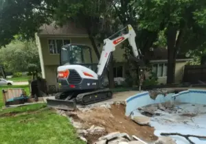 Mini excavator with a hydraulic breaker attachment demolishing an in-ground swimming pool in a residential backyard