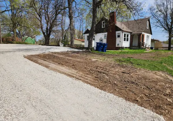 Freshly graded dirt driveway extension connecting to an existing gravel driveway beside a white house with trash bins