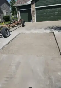 Square patch of compacted gravel base set into a concrete driveway in front of a green garage door, with an old trailer frame nearby