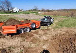 Orange dump trailer loaded with soil hitched to a pickup truck, parked in an open rural field near a farmhouse