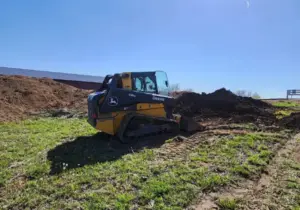 Yellow John Deere compact track loader parked in a mowed field next to piles of graded dirt under a clear sky